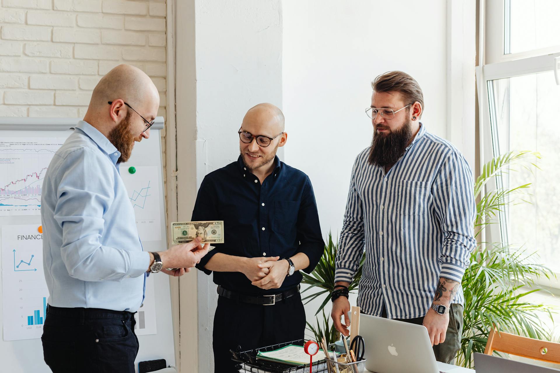 Three businessmen exchanging money in a modern office with financial charts.