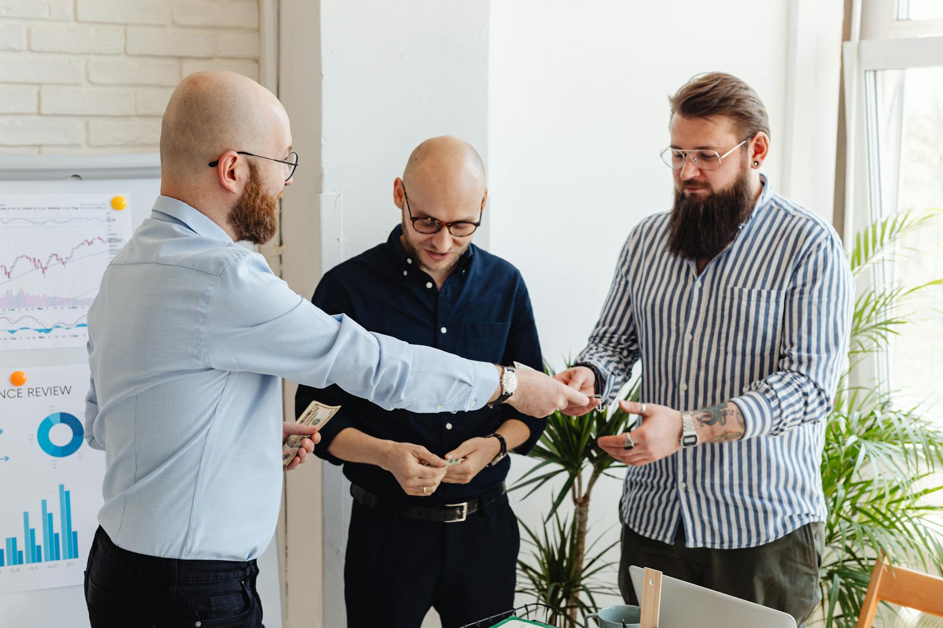 Three businessmen exchanging cash in a bright modern office setting with financial charts.