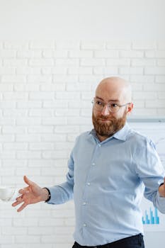 Bald businessman in blue shirt with beard and glasses gesturing in office.