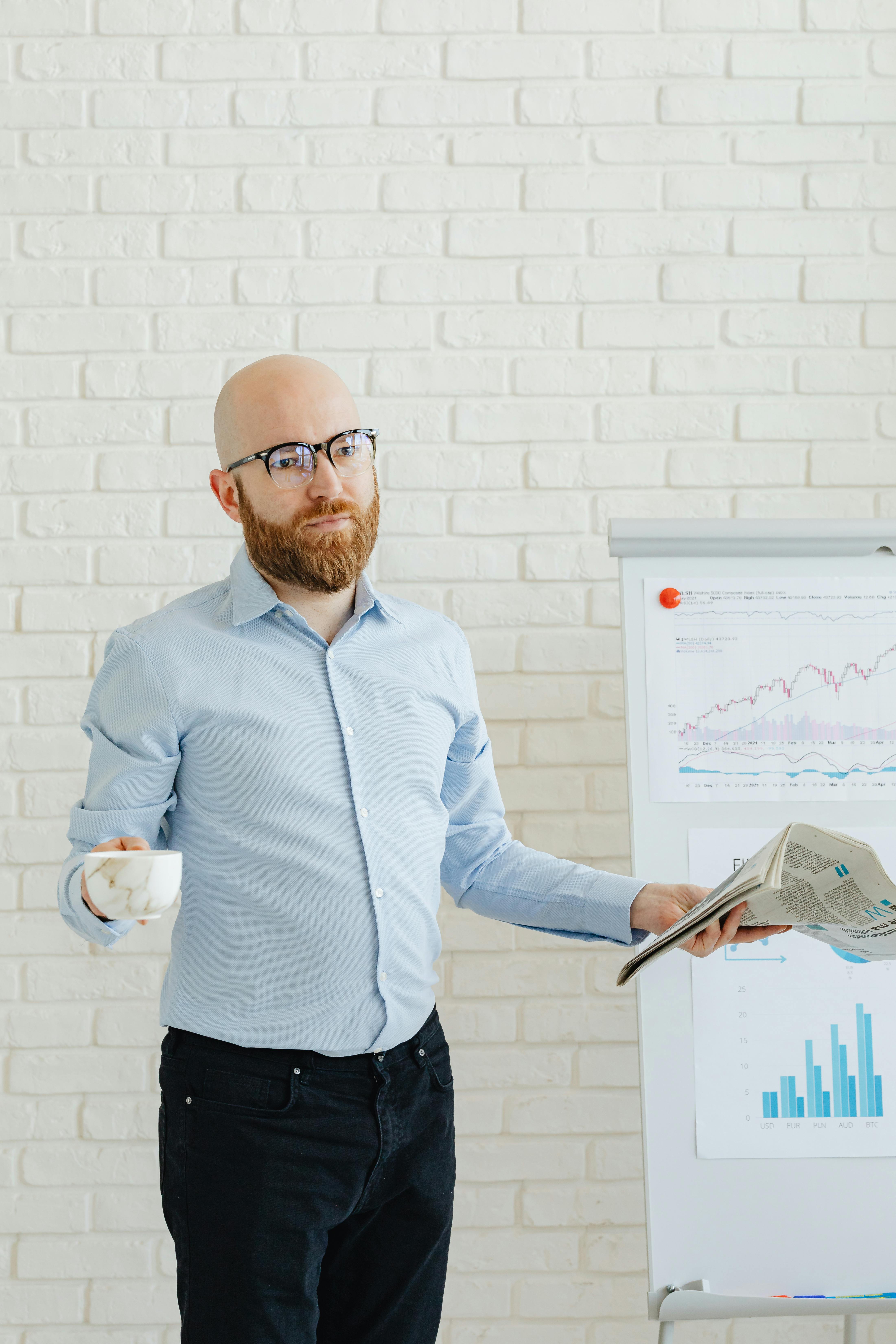 Bald man with beard holds a cup and newspaper while presenting on charts.