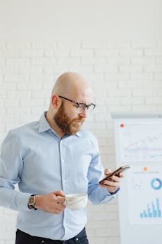 Bald man in corporate attire checks smartphone while holding a cup indoors.