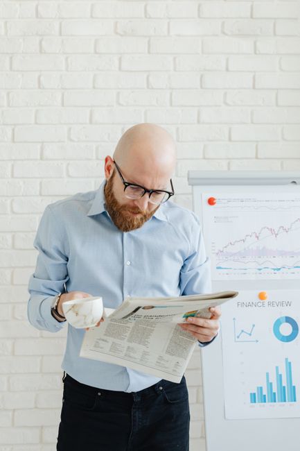 Bald man in glasses reading newspaper with coffee by presentation charts. Bald man in glasses reading newspaper with coffee by presentation charts.