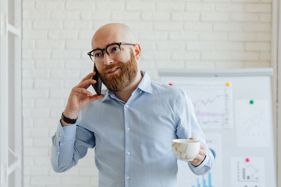 A bald man with eyeglasses standing indoors, talking on a smartphone while holding a ceramic cup.