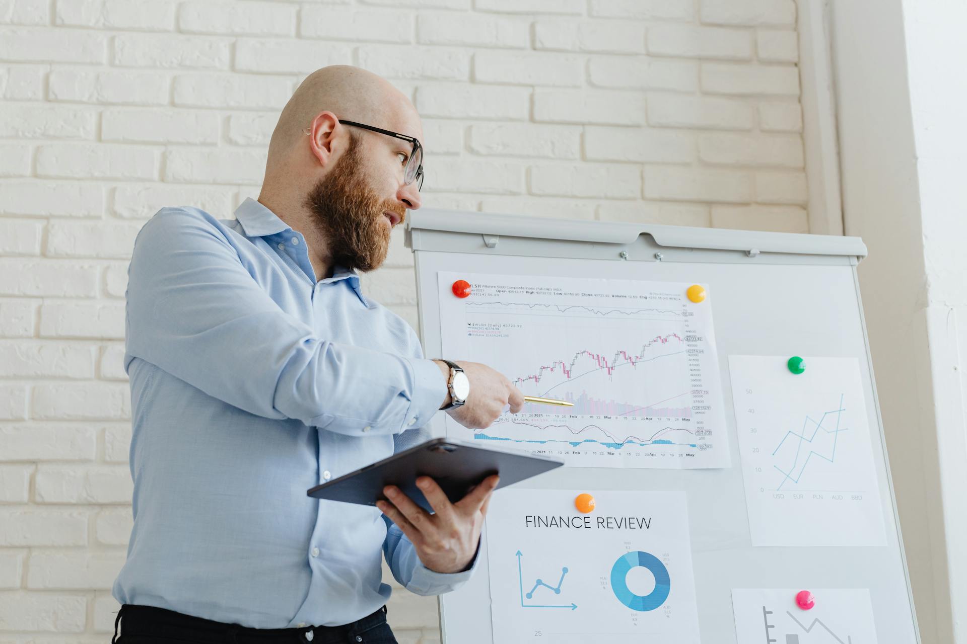 Man giving a business presentation with financial graphs on a whiteboard using a tablet for explanation.