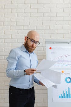 Bald man with eyeglasses reviewing financial papers in front of charts indoors.