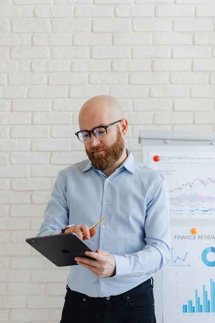 Bald man with beard reviews financial charts on a tablet in office.