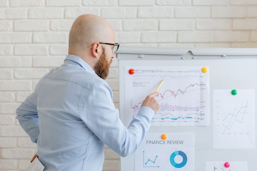 Bald businessman in office analyzes graphs on whiteboard, indicating finance trends.