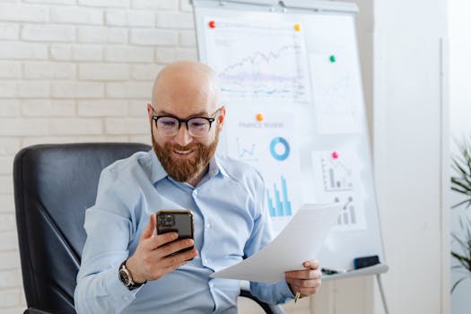 Smiling bald businessman checking smartphone while reviewing financial documents indoors.
