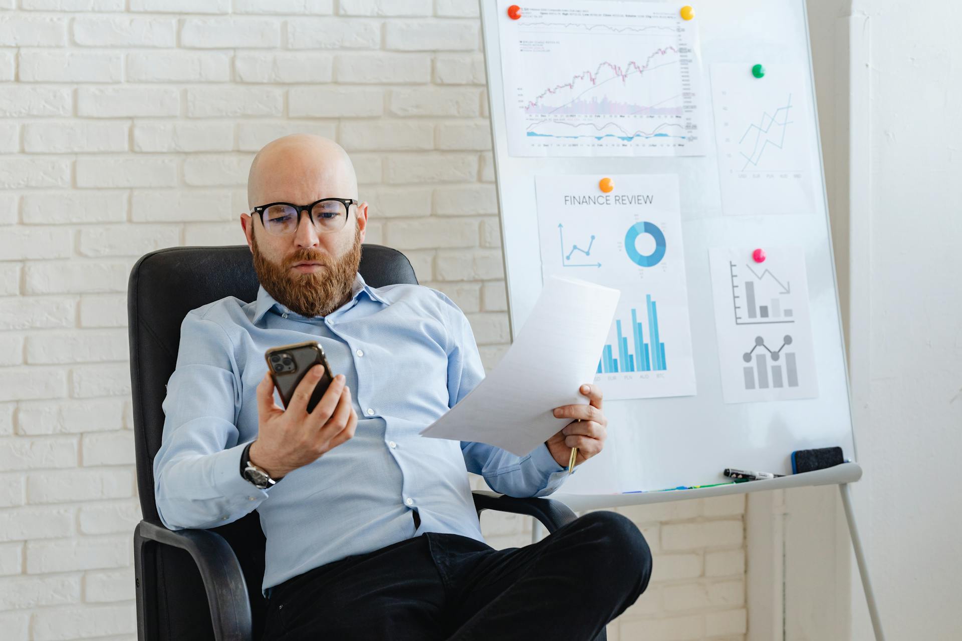 Bald businessman in corporate attire reviewing paperwork and smartphone data indoors.