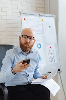 Bald businessman in office with smartphone, analyzing stock market charts on papers.