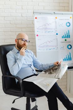 Bald businessman sitting with newspaper, analyzing financial charts in modern office.