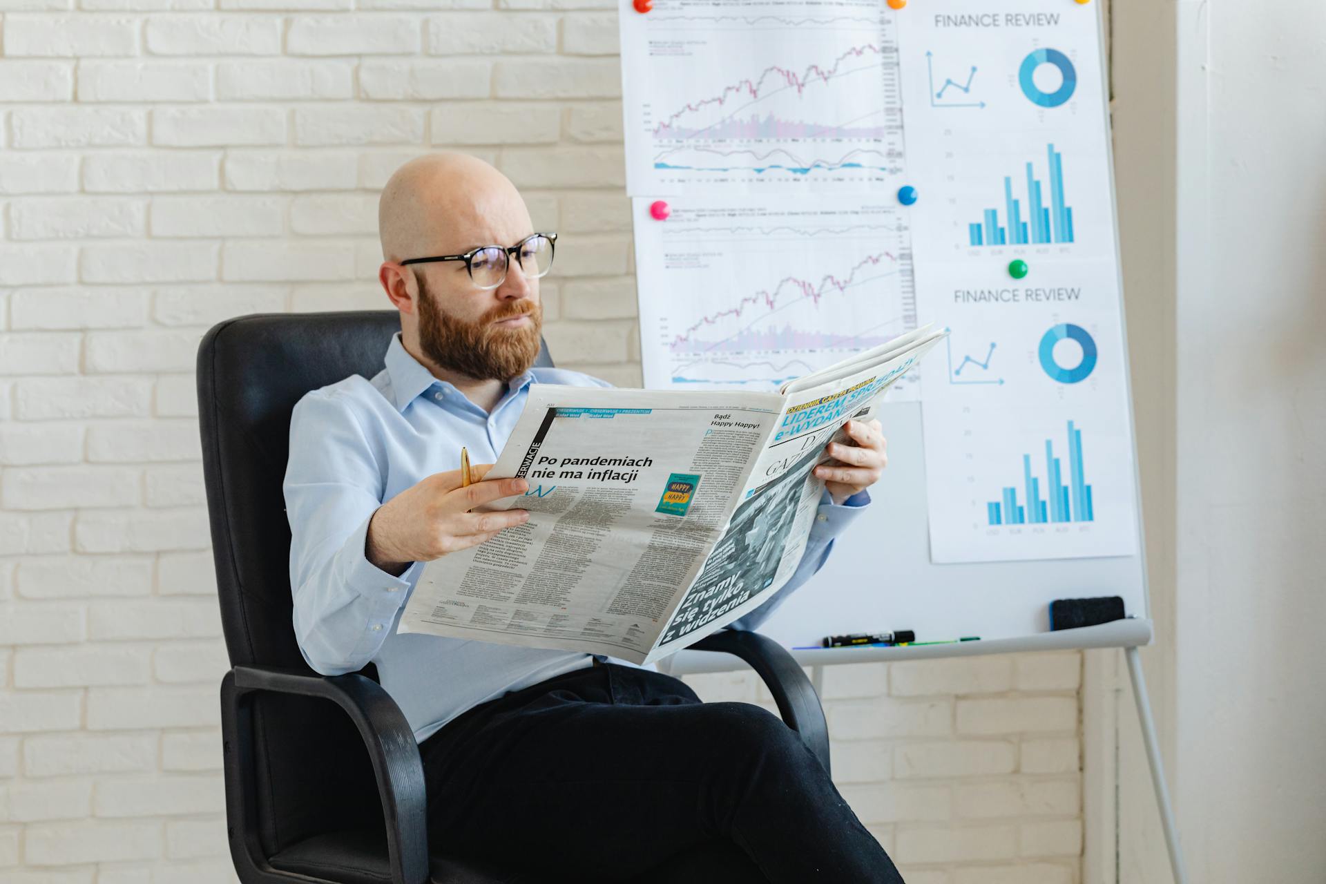 Bald man with beard reading newspaper in office with finance charts on whiteboard.