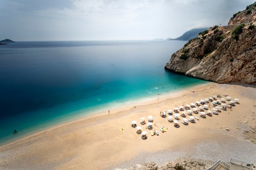 Aerial view of picturesque scenery of sandy beach with umbrellas and lounge chairs rocky cliff and turquoise ocean