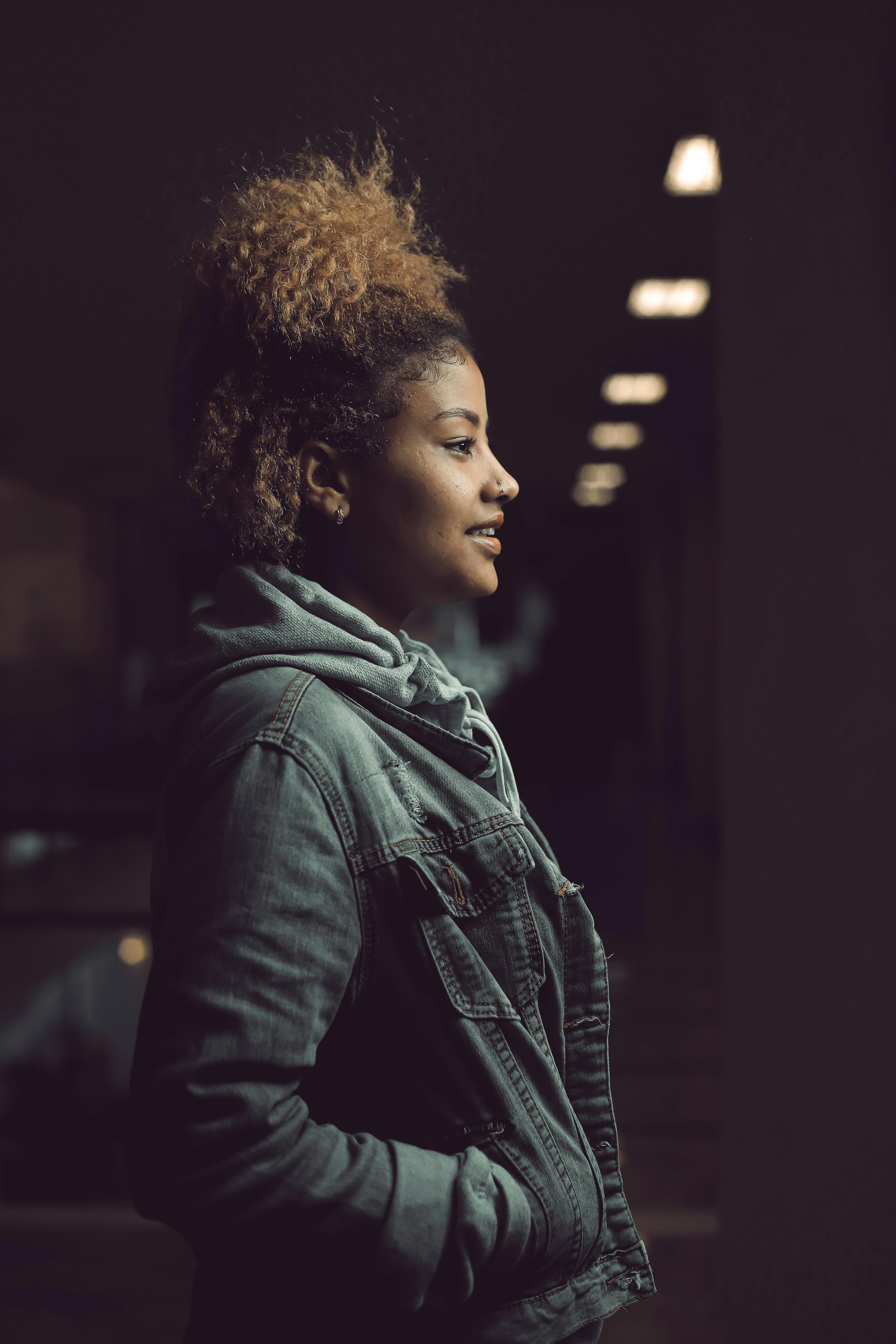Side view of a woman in a denim jacket with curly hair against a dark background.