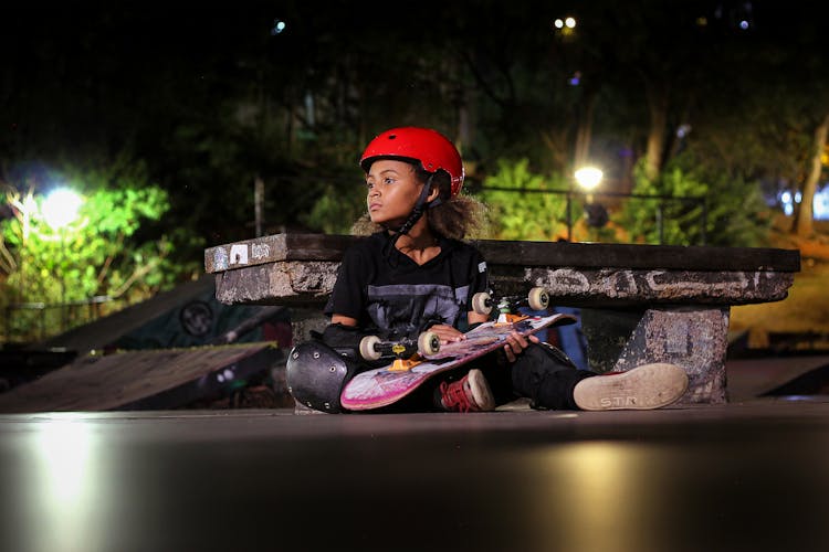 A Kid Wearing Red Helmet Sitting Beside A Concrete Bench Holding A Skateboard