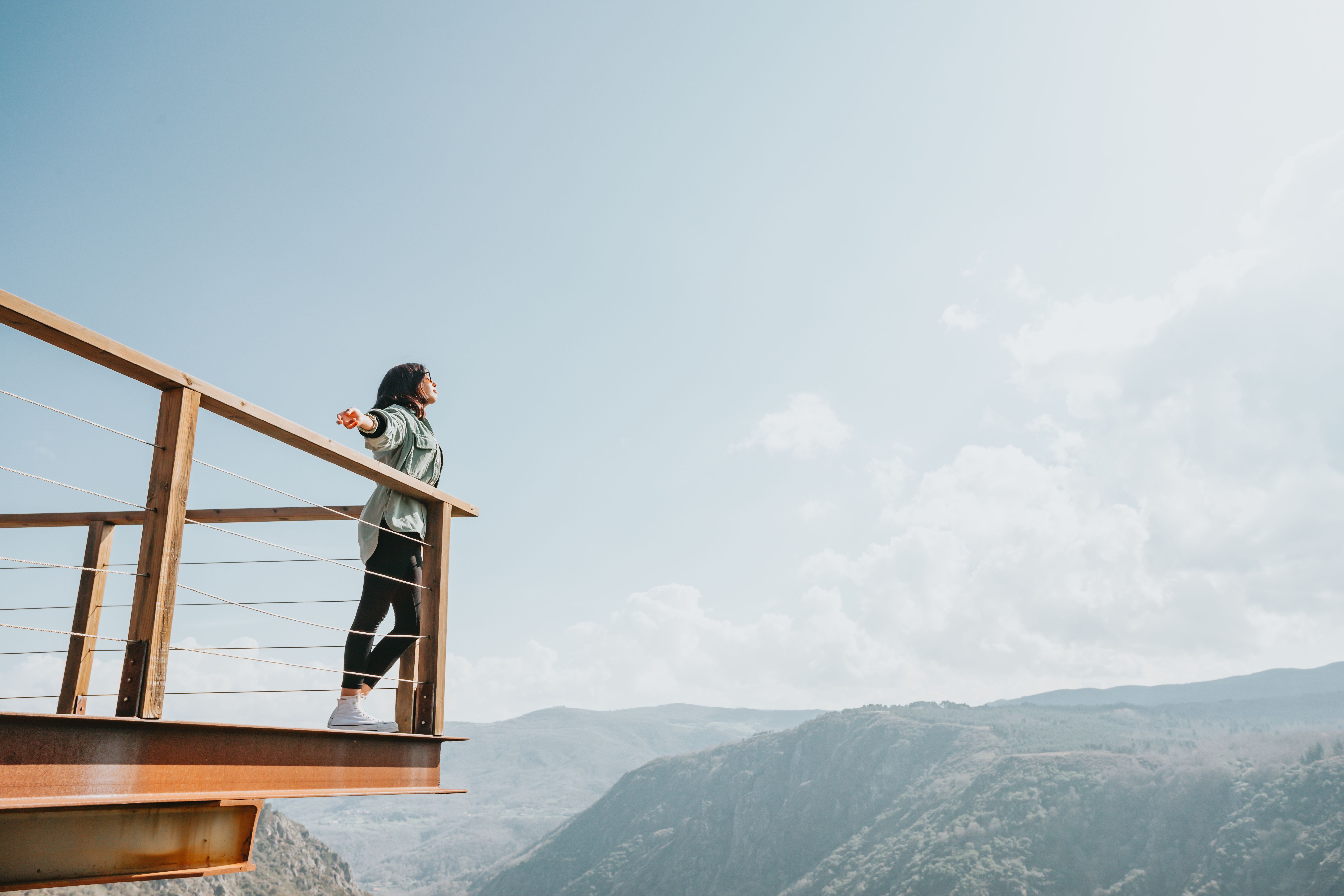 A woman relishes the adventurous mountain view from a scenic deck under a bright blue sky.