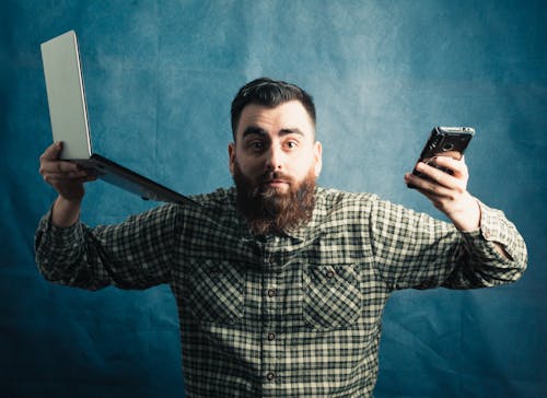Bearded Man Holding Cellphone and Laptop on Blue Background