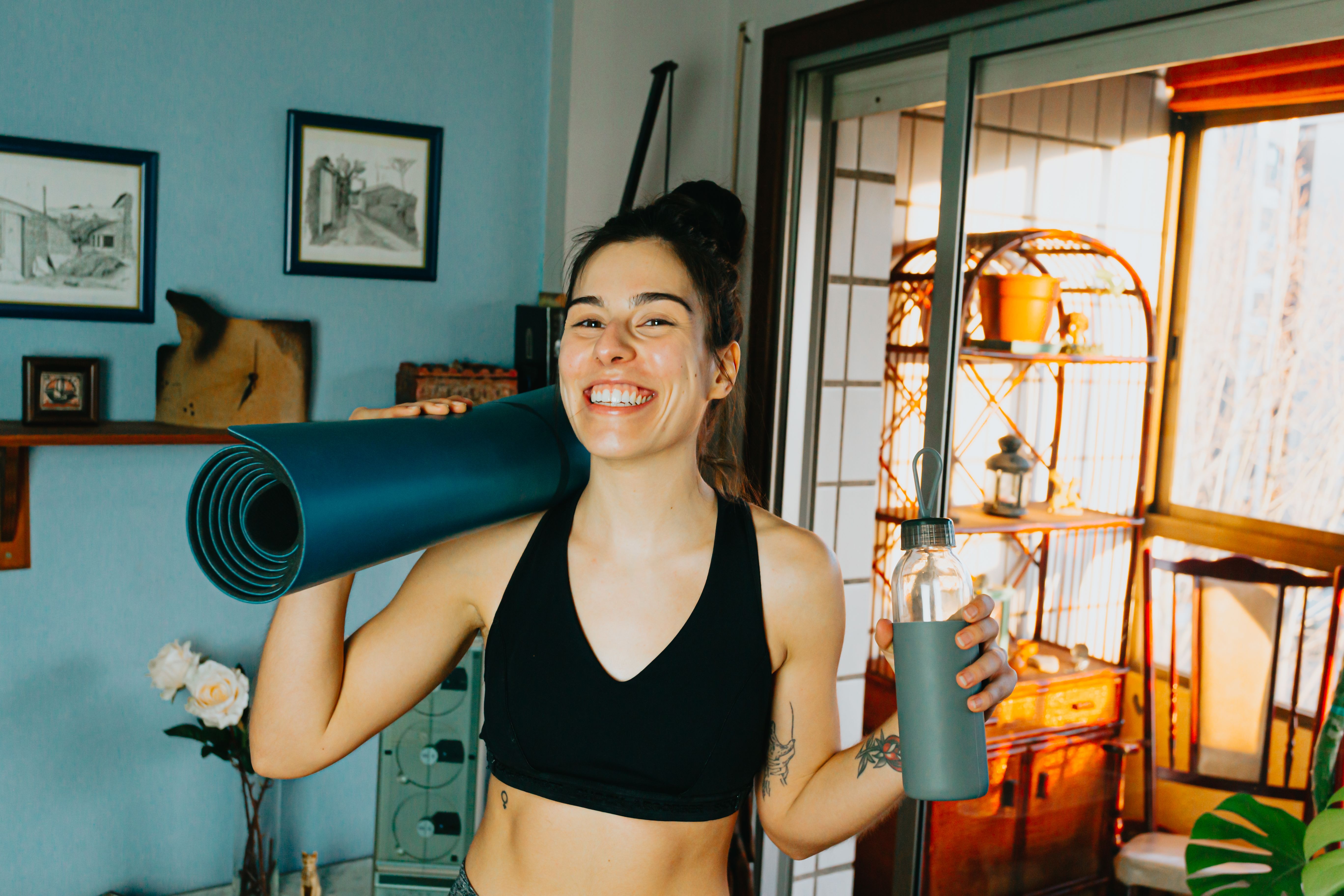Cheerful woman with yoga mat and bottle ready for home workout in cozy room.