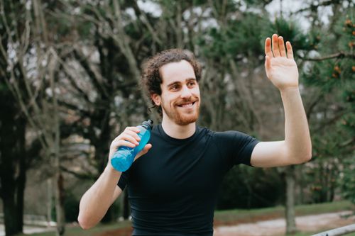 Free Man in a park waving while holding a water bottle, expressing joy and friendliness. Stock Photo
