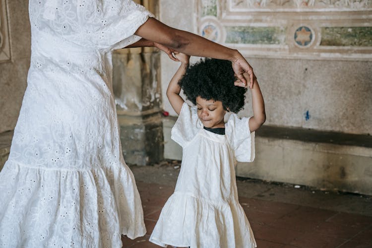 A Little Girl In A White Dress Dancing With Her Mom