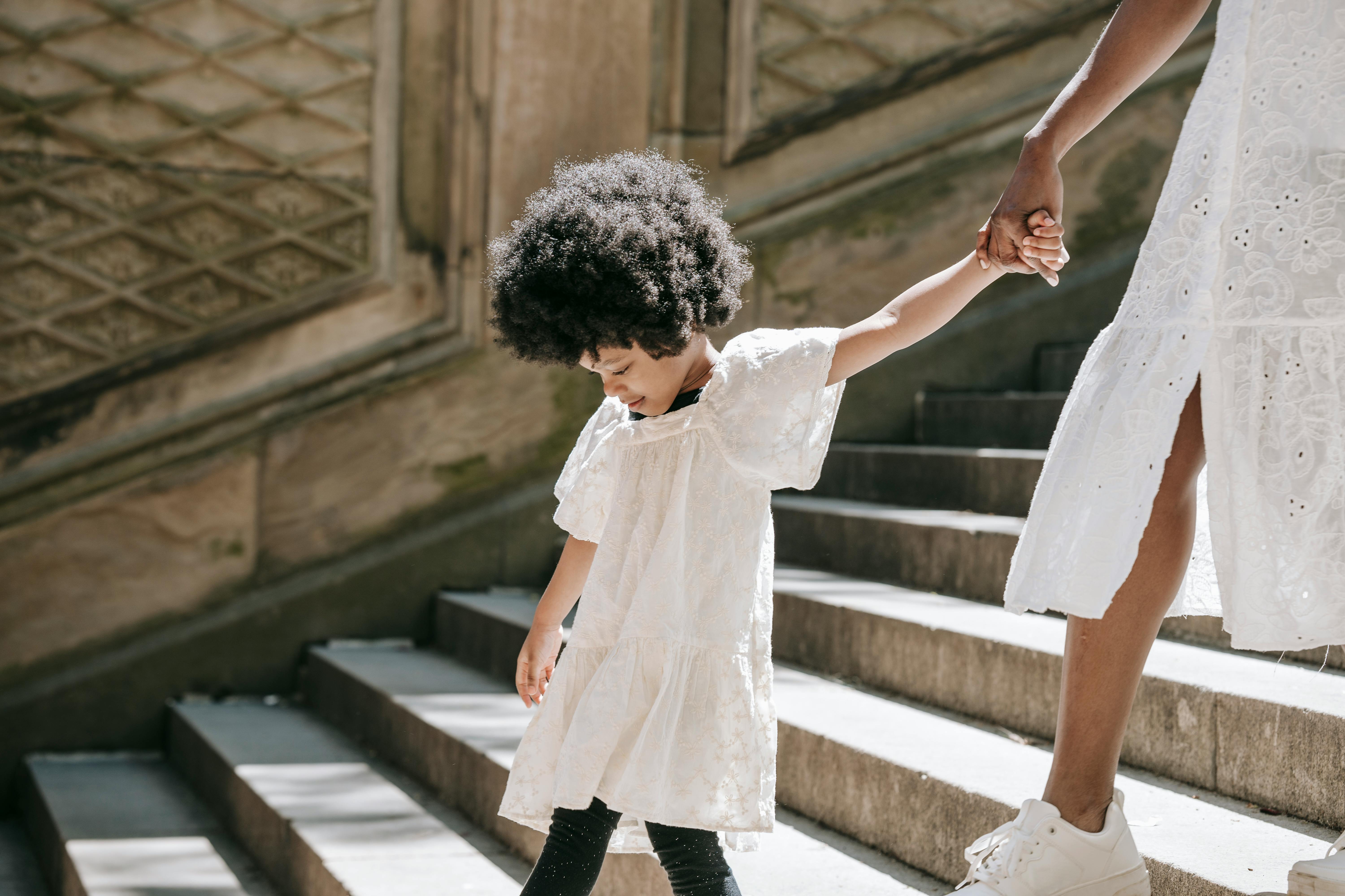 A young girl in a white dress with afro hair holding hands and walking down stone stairs outdoors.
