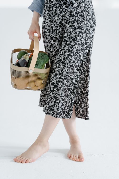 Free Barefoot woman in floral skirt holding a basket of fresh vegetables indoors. Stock Photo