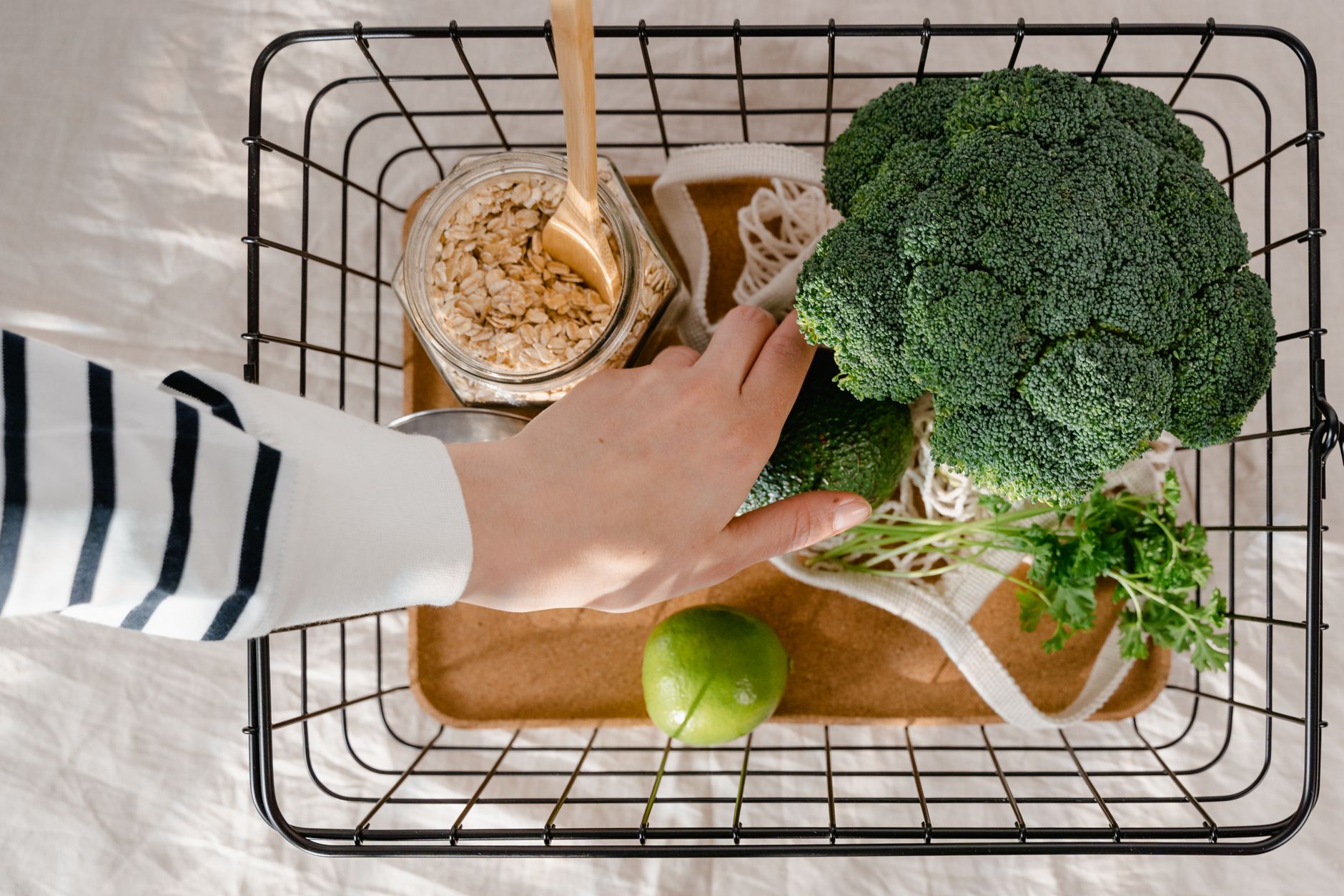 Overhead view of a basket with broccoli, lime, oatmeal jar, and a hand reaching in. Fresh and healthy concept.