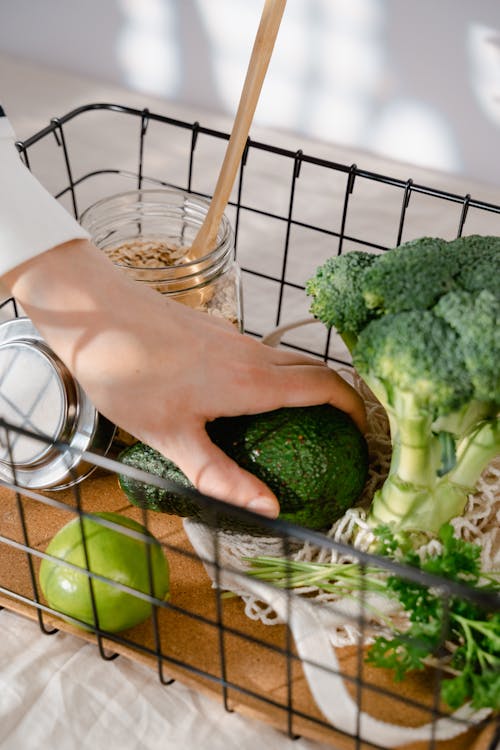 A Person Holding a Green Avocado Beside a Broccoli and Lime