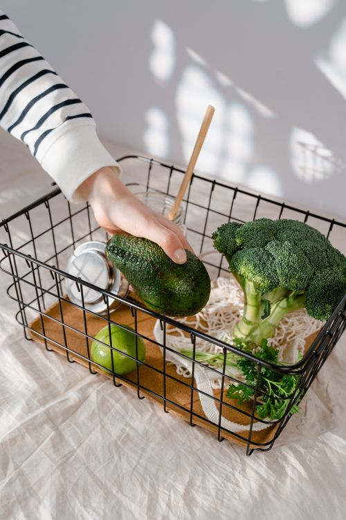 Free Person's hand placing an avocado in a basket with broccoli and lime, under natural light. Stock Photo