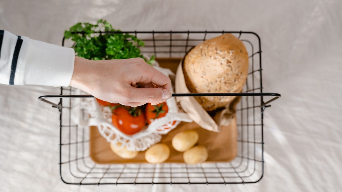 Top view of a hand holding a basket with bread, potatoes, tomatoes, and greens.