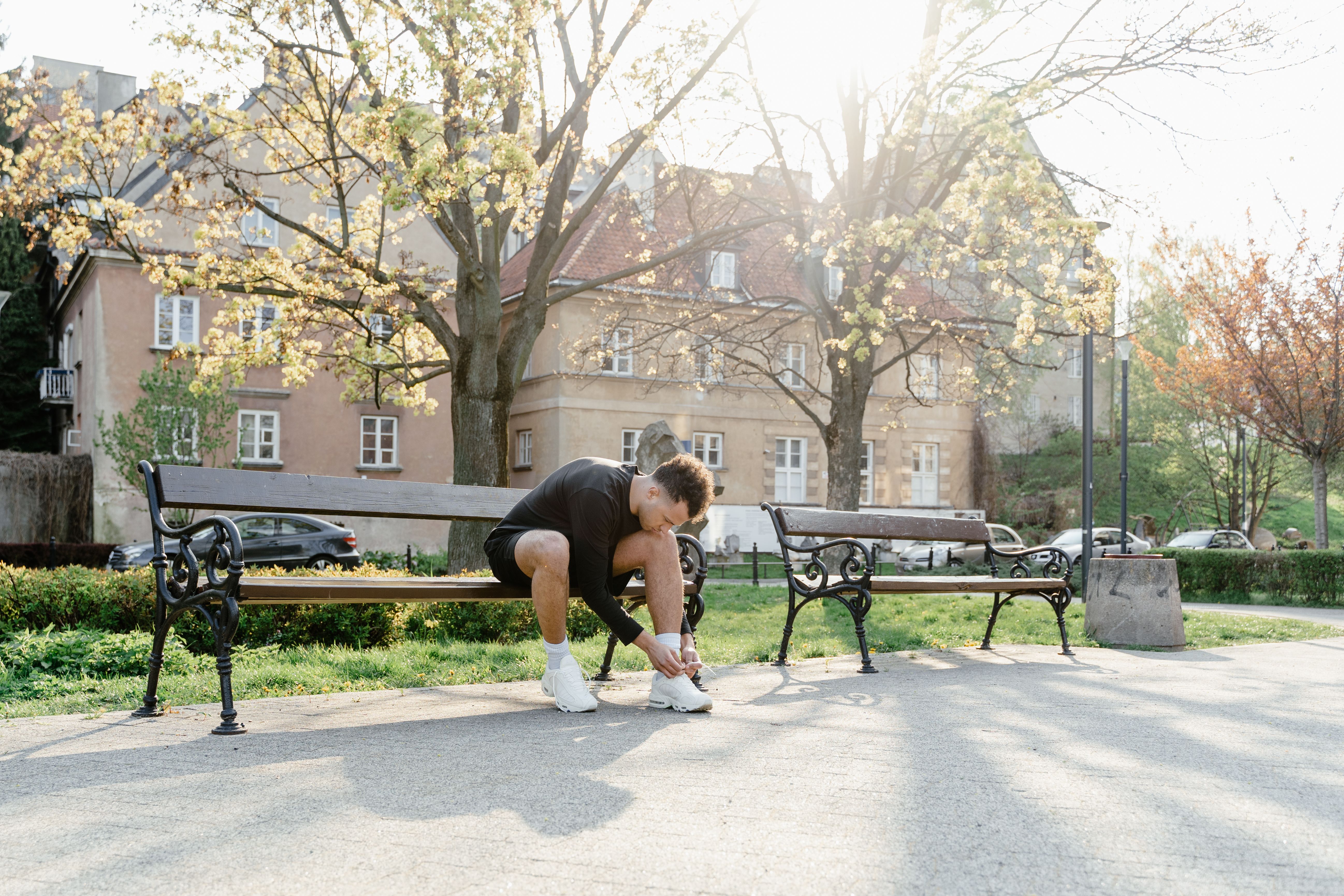 A man in sportswear ties his sneakers on a bench in a sunny park setting, ready for exercise.