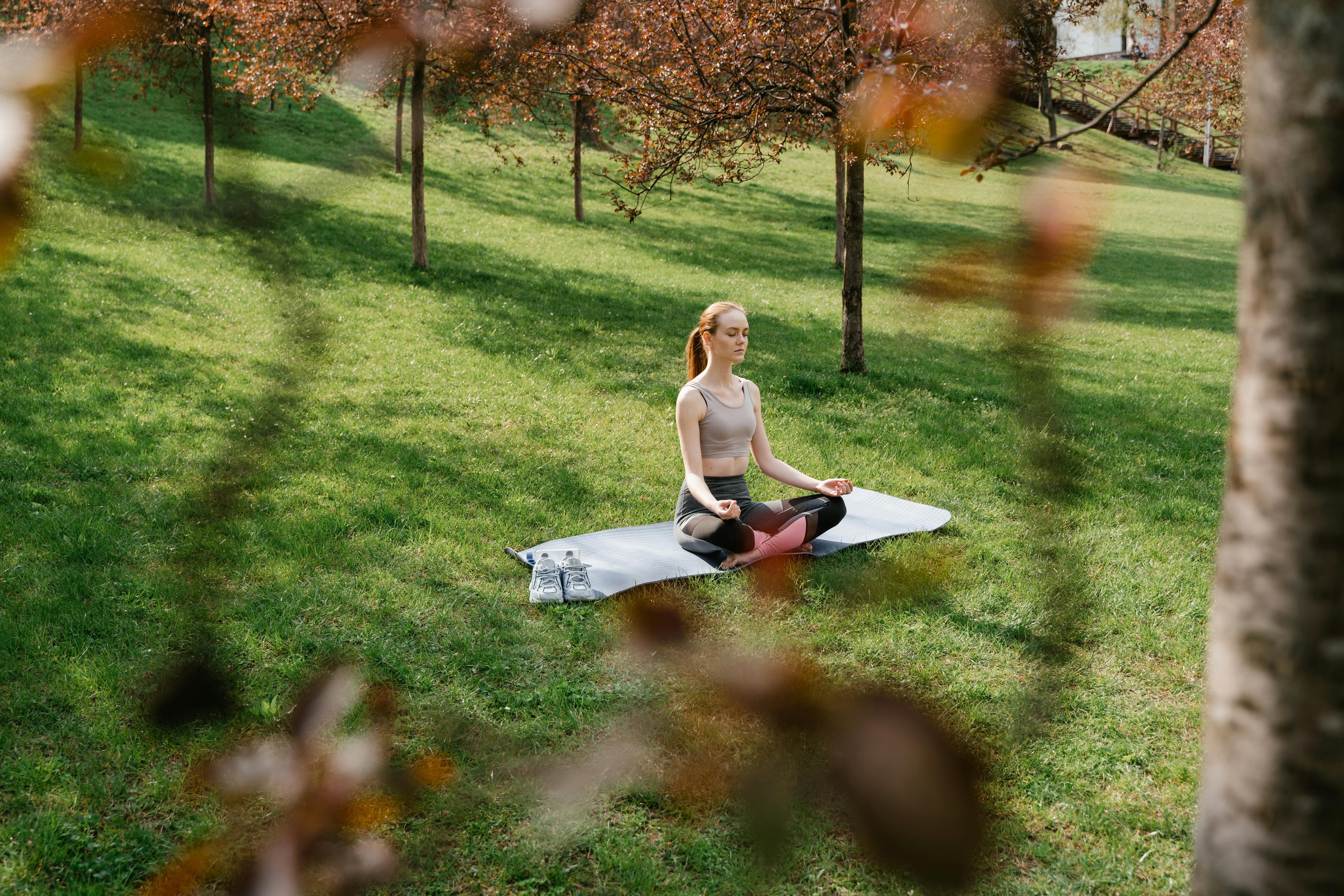 A woman meditating on a yoga mat in a sunny park surrounded by trees.