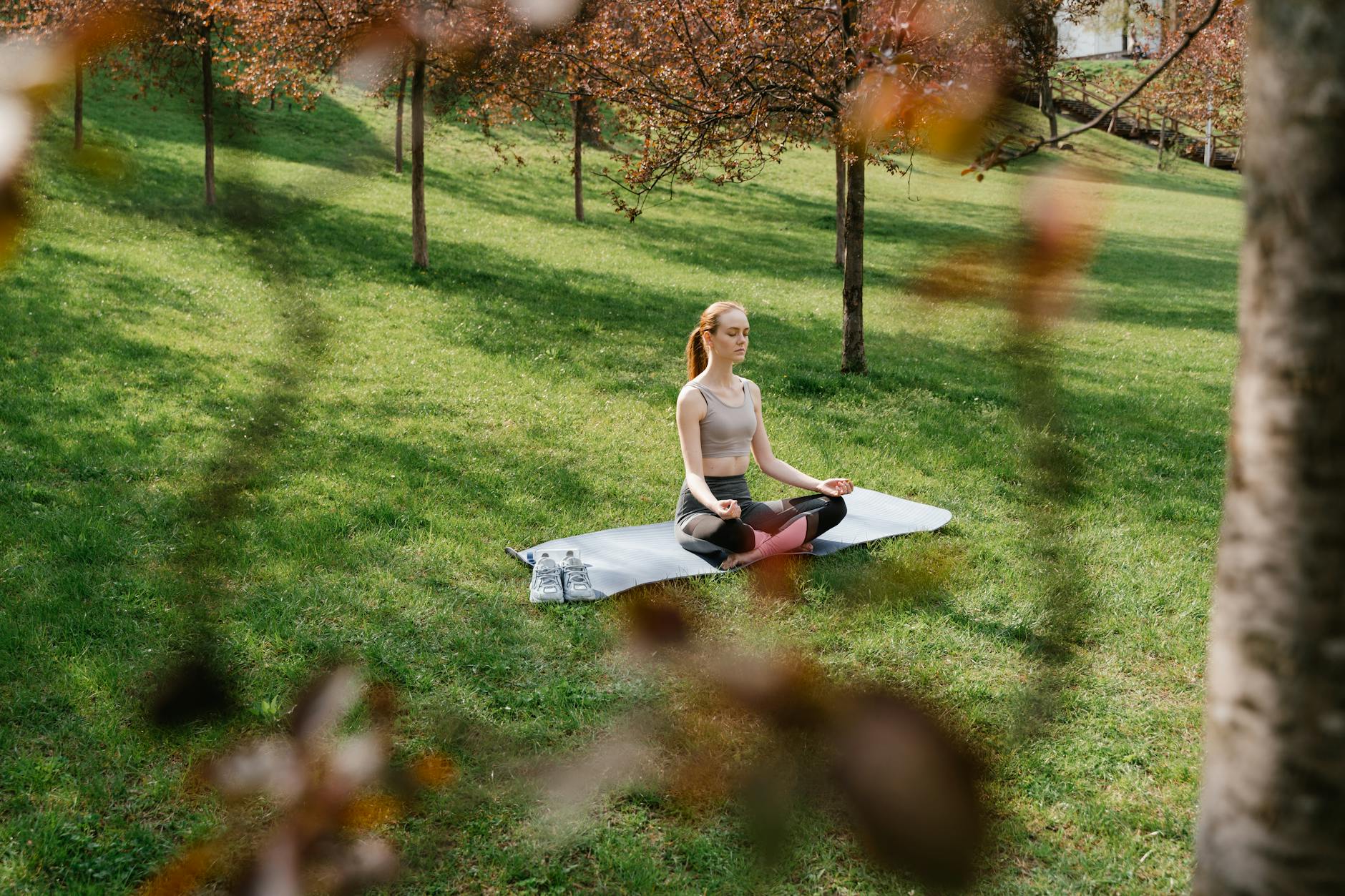 A woman meditating on a yoga mat in a sunny park surrounded by trees.