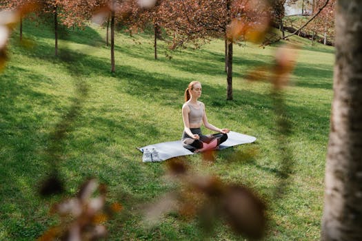 A woman meditating on a yoga mat in a sunny park surrounded by trees.