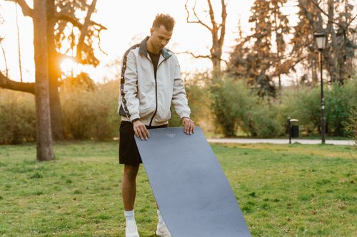 Free A man prepares a yoga mat outdoors in a vibrant park setting at sunset. Stock Photo