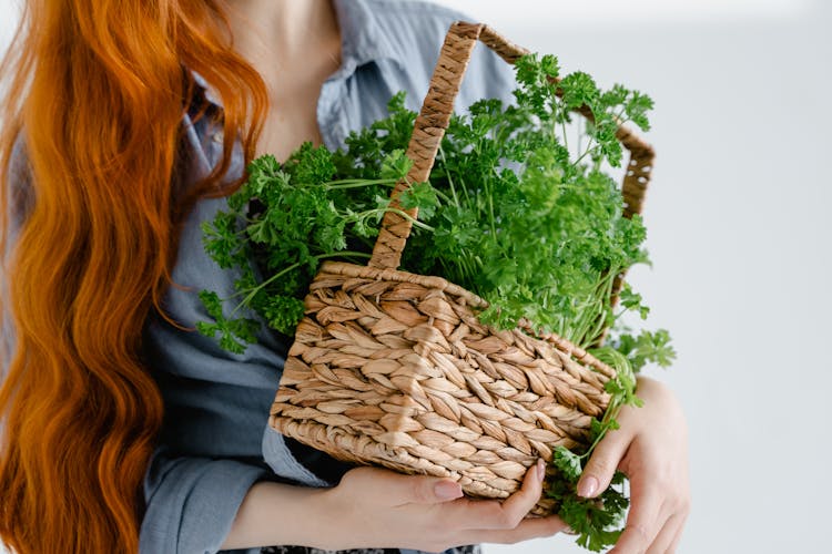 A Person With Long Hair  Holding Woven Basket With A Bunch Of Parsley