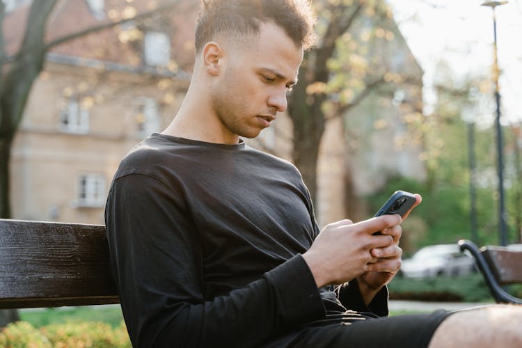 A Man In Black Long Sleeve Shirt Sitting On Bench Holding A Cellphone