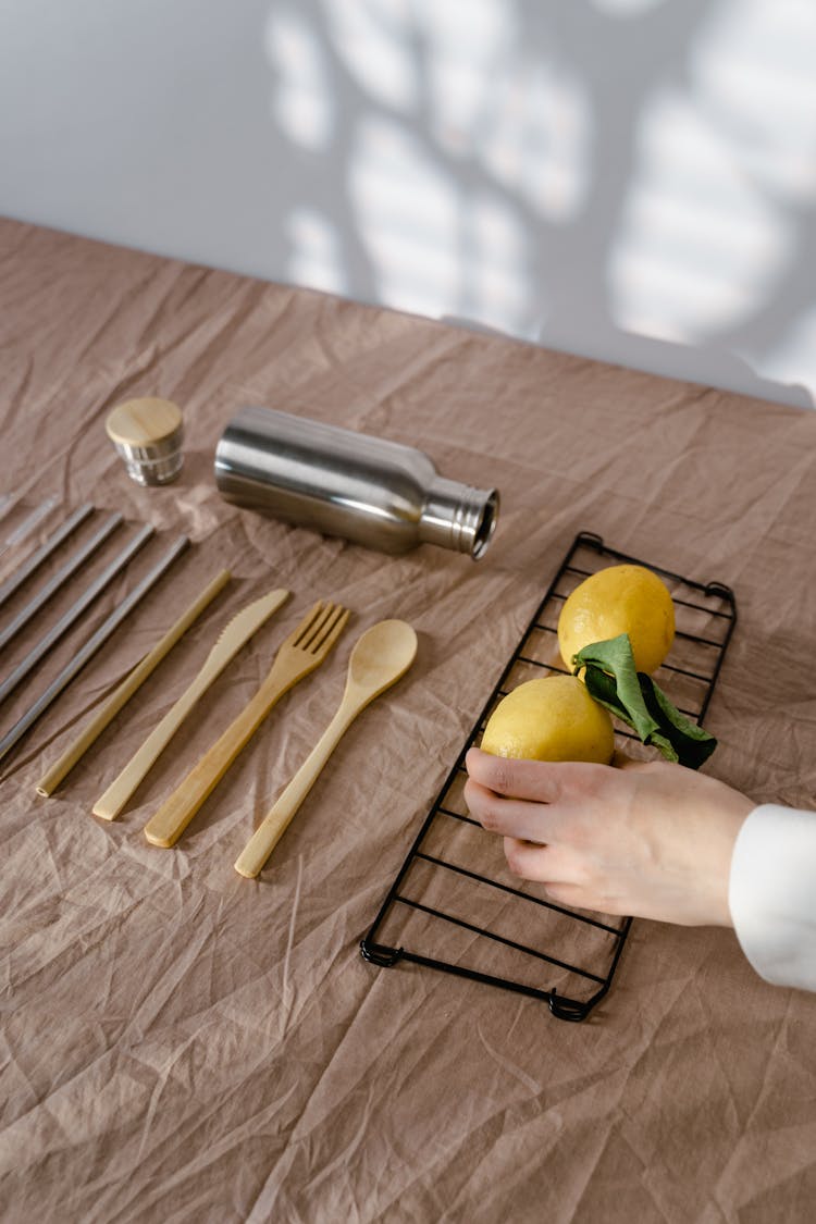 Person Holding A Lemon On Table Beside A Stainless Steel Vacuum Flask And Wooden Utensils