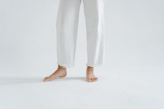 Close-up of barefoot legs in white pants against a minimalist white background.