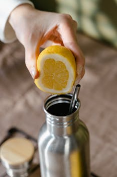 Close-up of a hand squeezing a lemon into a stainless steel water bottle with a lid.