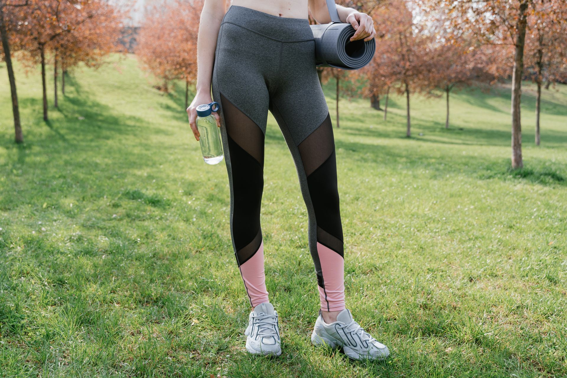Fit woman outdoors with yoga mat and water bottle ready for exercise.