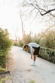 A man in sportswear bends over, stretching along a sunlit park path surrounded by trees.