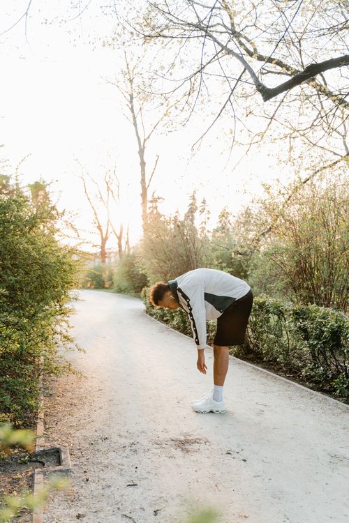 Free A man in sportswear bends over, stretching along a sunlit park path surrounded by trees. Stock Photo