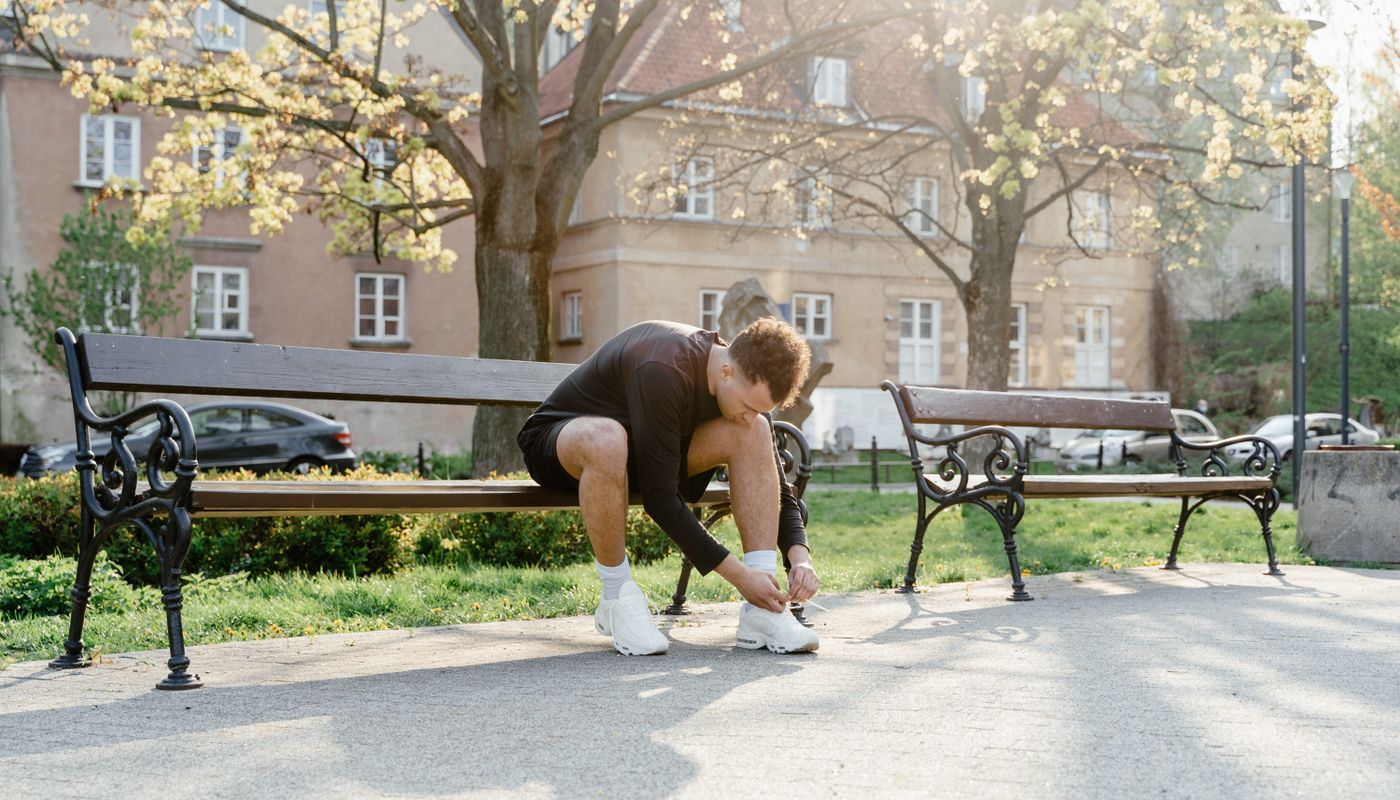A runner sitting on a park bench in morning light, bending down to tie a shoelace