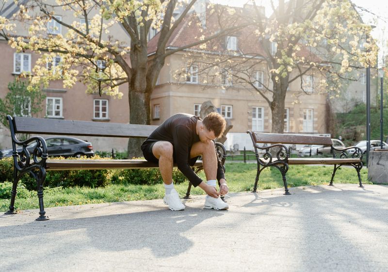A runner sitting on a park bench in morning light, bending down to tie a shoelace