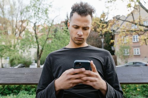 Free Casual young man sitting on a bench, texting on his smartphone in a park setting. Stock Photo