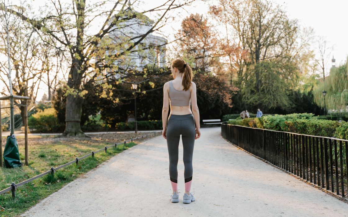 Woman in activewear walking along a sunny tree-lined park path, seen from behind