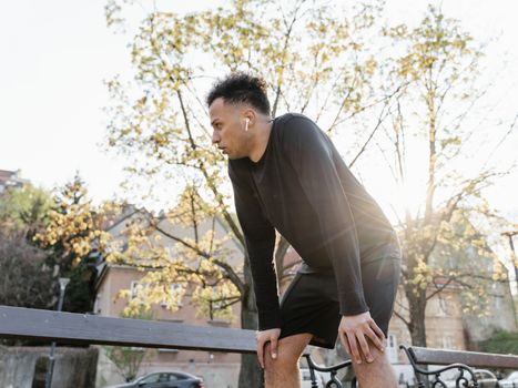 Man in athletic gear pauses during a run, wearing earbuds in a sunny park.