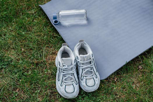 Pair of sneakers with a yoga mat and water bottle on grass, ideal for outdoor workouts.