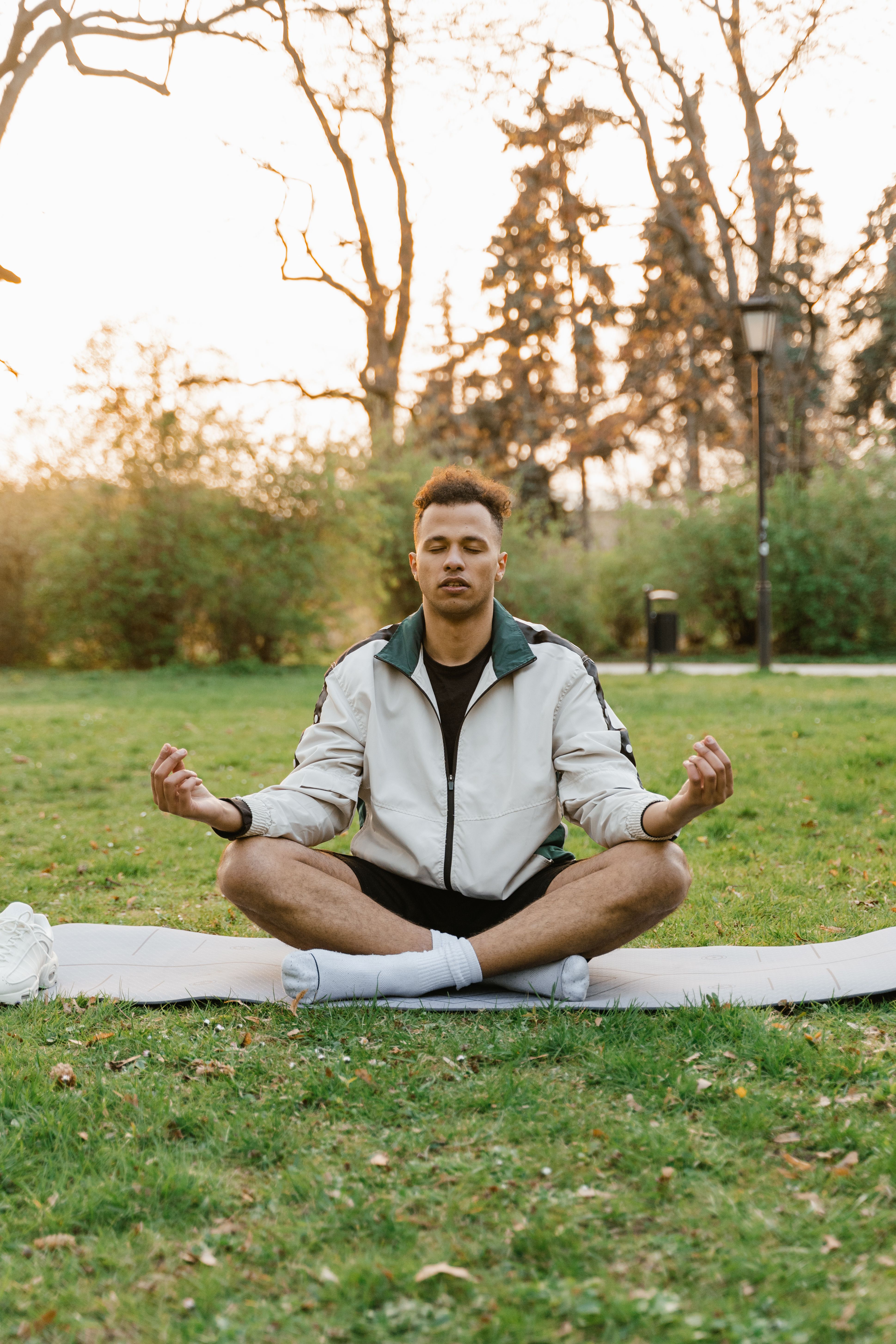 Young man in lotus pose meditating outdoors on a yoga mat at sunset, promoting relaxation and wellness.